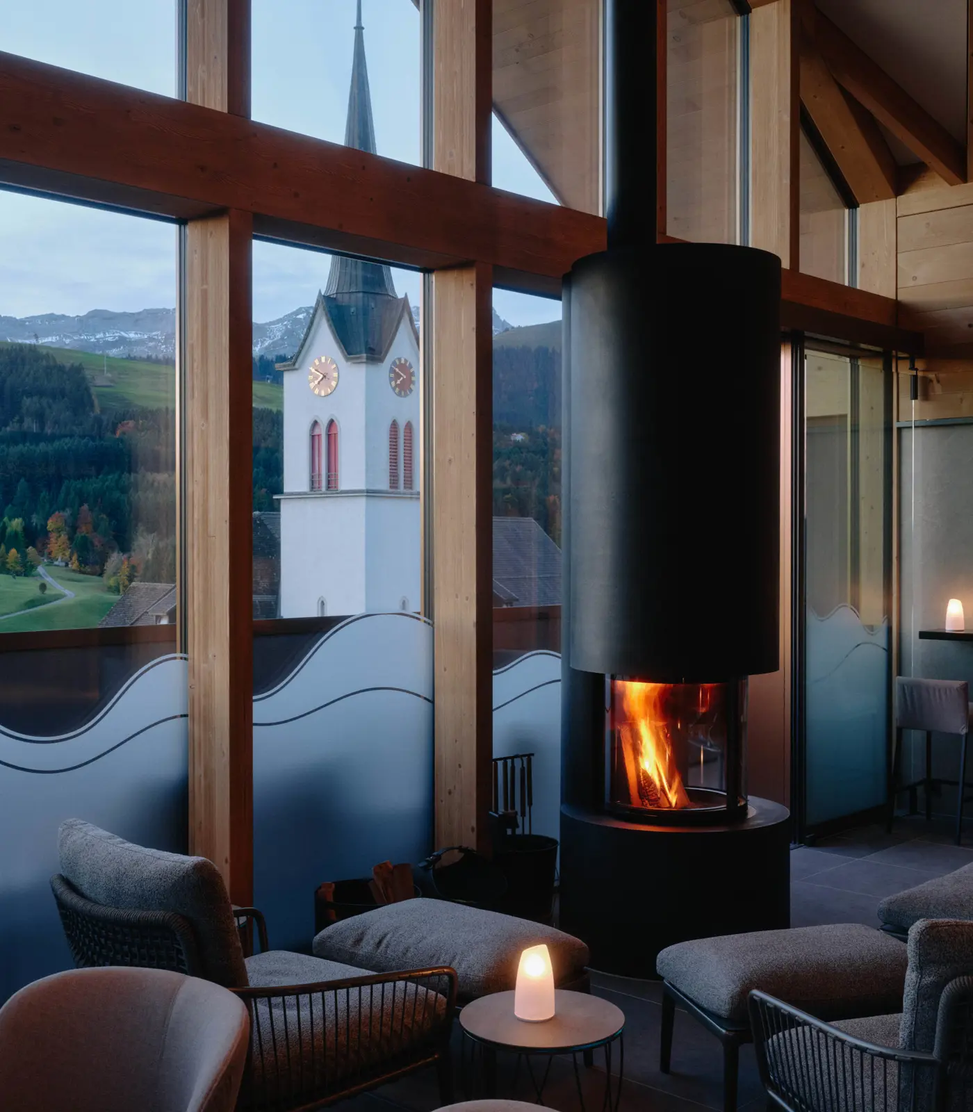 A fireside lounge at Appenzeller Huus, with upholstered chairs drawn around the fire and tall windows looking out over Gonten